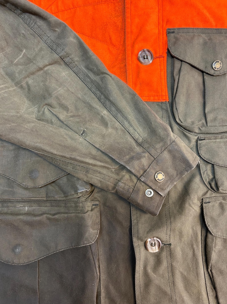 May include: A brown and orange waxed cotton hunting jacket with multiple pockets and button closures. The jacket has a  "Filson" logo on the sleeve.
