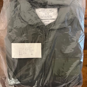May include: An unworn, olive green coverall jacket, sealed in a clear plastic bag. The jacket features a zip closure and two front pockets. A label on the jacket reads "44 R" and includes other text. The jacket is new.