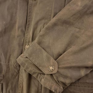 Peut inclure: Gros plan sur une veste marron avec un poignet boutonn&eacute; et une fermeture &eacute;clair. La veste comporte plusieurs boutons avec un motif circulaire. Le tissu a un aspect l&eacute;g&egrave;rement us&eacute;.