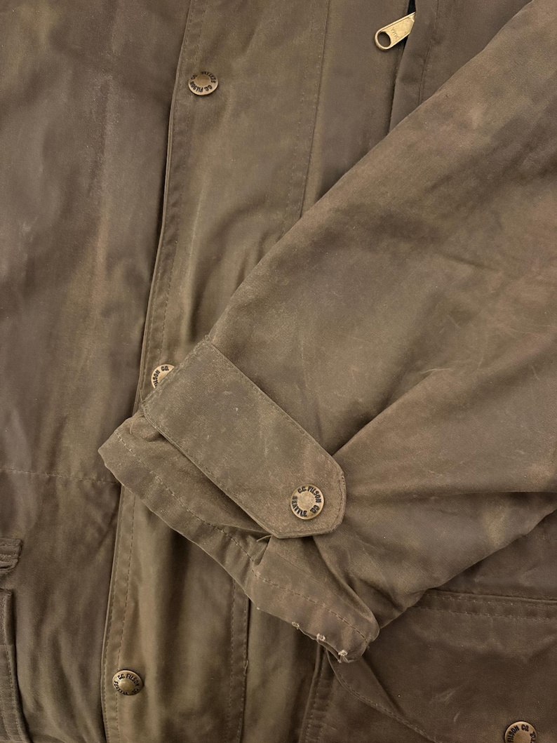 Peut inclure: Gros plan sur une veste marron avec un poignet boutonn&eacute; et une fermeture &eacute;clair. La veste comporte plusieurs boutons avec un motif circulaire. Le tissu a un aspect l&eacute;g&egrave;rement us&eacute;.