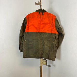 May include: A brown and orange hunting jacket with a zipper closure and multiple pockets. The jacket is hanging on a wooden stand.