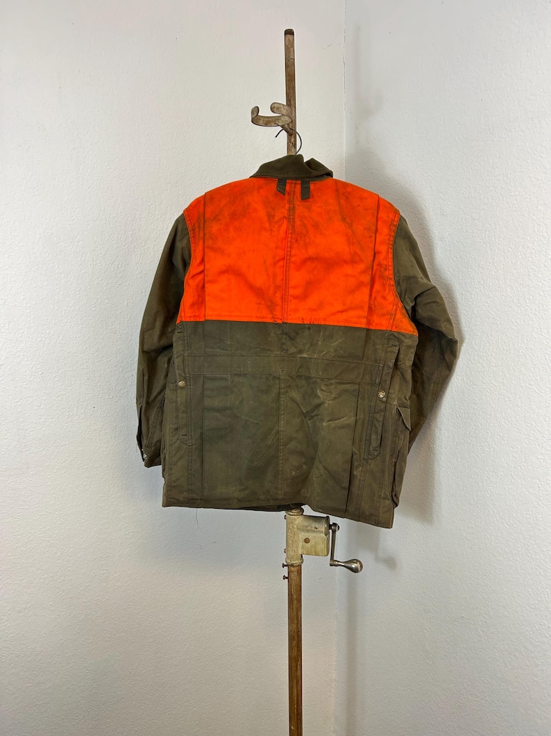 May include: A brown and orange hunting jacket with a zipper closure and multiple pockets. The jacket is hanging on a wooden stand.