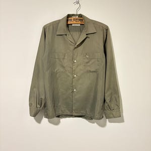 Puede incluir: Camisa de manga larga verde oliva con dos bolsillos en el pecho y cierre de botones. La camisa está colgada de una percha de madera vintage con el texto "ROBE CLOTHING". La camisa tiene un estilo clásico e informal.