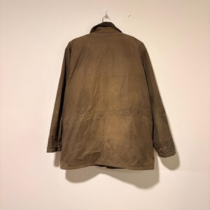 Peut inclure: Veste marron avec un col marron fonc&eacute; suspendue &agrave; un crochet en m&eacute;tal. La veste a un devant boutonn&eacute;, des manches longues et une taille &agrave; cordon. La veste est faite d'un mat&eacute;riau durable et r&eacute;sistant &agrave; l'eau.