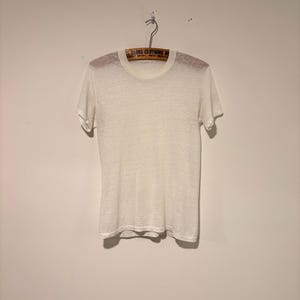May include: A semi-sheer, off-white short-sleeve t-shirt with a crew neckline. The garment is displayed on a wooden hanger that features the text GLOBE CLOTHING HOUSE printed in black. The fabric has a visible, fine-knit texture throughout.