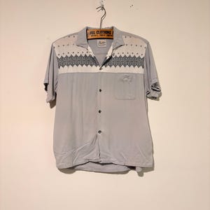 May include: A light gray short-sleeve button-down shirt featuring a white yoke with a dark gray geometric diamond pattern across the chest and shoulders. The shirt includes a collar, a single chest pocket, and dark buttons down the front.