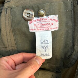 May include: A green cotton jacket with a brown button and a label that reads "A Genuine Filson Garment" and "C-C-Filson Co. Seattle" with the tagline "Might as well have the best". The label also includes the style number 463, 9406 2677, size S, and "100% Cotton Care on reverse".