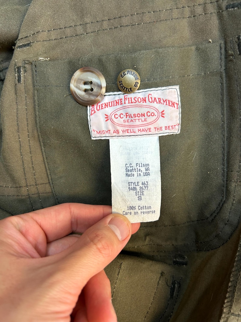 May include: A green cotton jacket with a brown button and a label that reads "A Genuine Filson Garment" and "C-C-Filson Co. Seattle" with the tagline "Might as well have the best". The label also includes the style number 463, 9406 2677, size S, and "100% Cotton Care on reverse".