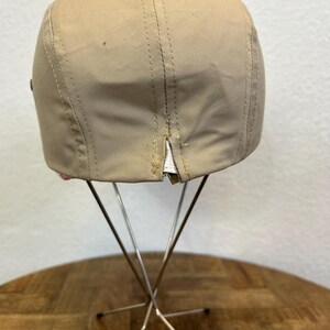 May include: A tan baseball cap with a curved brim, displayed on a metal stand. The cap has visible stitching and a small tear near the back. The cap is made of a smooth material.