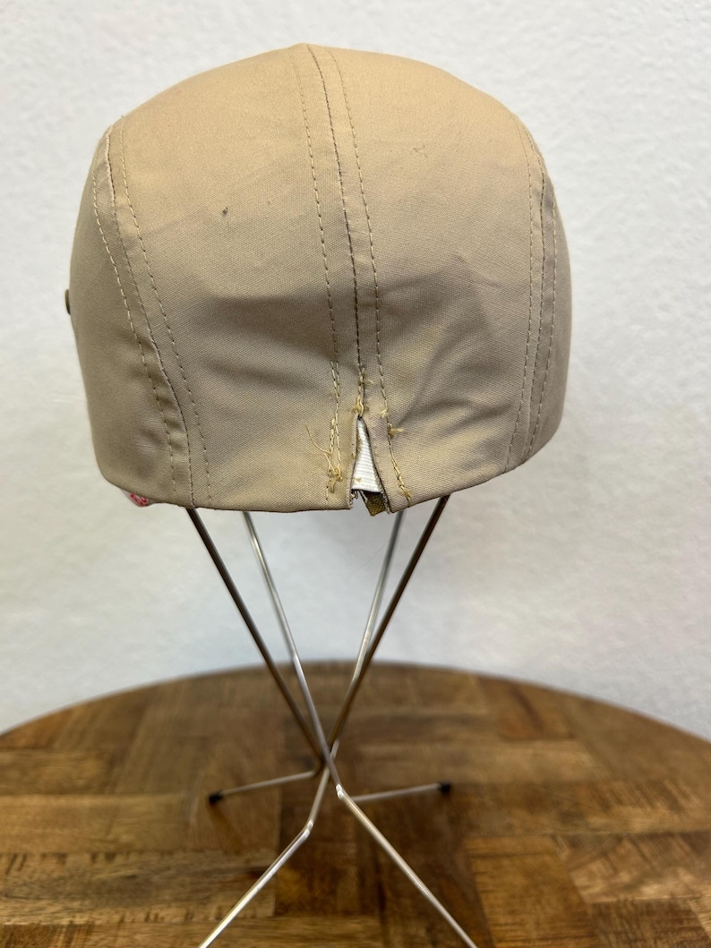 May include: A tan baseball cap with a curved brim, displayed on a metal stand. The cap has visible stitching and a small tear near the back. The cap is made of a smooth material.