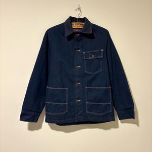 May include: A dark blue denim jacket with a brown collar and three front pockets. The jacket has a button-up closure and red stitching details. The label on the collar reads "BE CLOTHING".