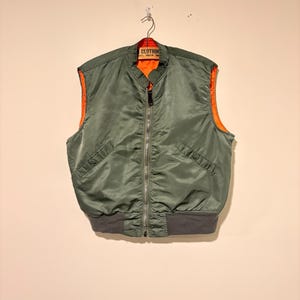 Puede incluir: Un chaleco tipo bomber sin mangas de color verde oliva con forro interior naranja brillante, cremallera frontal plateada, dos bolsillos laterales inclinados con botones a presión y un dobladillo de punto acanalado gris oscuro.