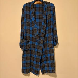 May include: A long, open-front plaid jacket in shades of blue, black, and grey hangs on a wooden hanger. The jacket features a classic plaid pattern with rolled-up sleeves and two front pockets. The fabric has a subtle sheen.