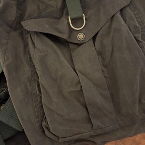 May include: A close-up of a dark olive green backpack. It features a large front pocket and a smaller pocket on top. The backpack has a green strap with a metal D-ring and a button closure. The material appears to be waxed canvas.
