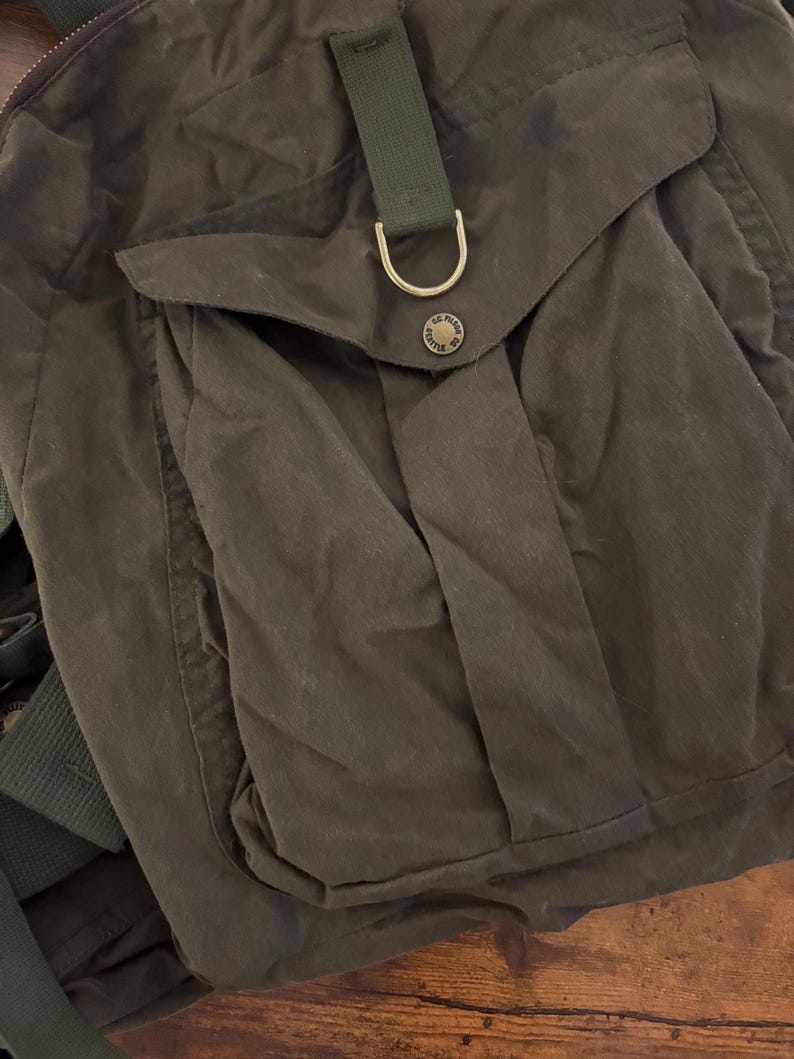 May include: A close-up of a dark olive green backpack. It features a large front pocket and a smaller pocket on top. The backpack has a green strap with a metal D-ring and a button closure. The material appears to be waxed canvas.