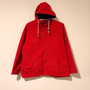 May include: A bright red hooded jacket with a blue interior lining and white drawstrings. The jacket has a button-up front, two front pockets, and a drawstring hood. A classic outerwear piece.
