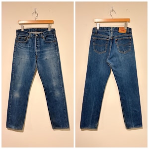 May include: A pair of blue denim jeans shown from the front and back. The jeans feature a button fly, five pockets, and a tan leather patch on the back waistband. The denim has a medium wash with subtle fading and whiskering throughout the fabric.