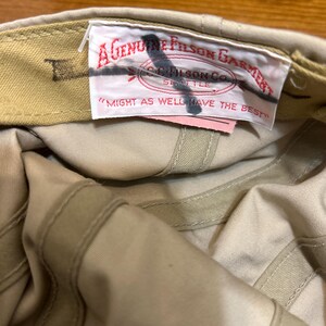 May include: Tan-colored cap with a label that reads "A Genuine Filson Garment" and "Might as well have the best." The cap is made of a durable fabric and features a classic design.