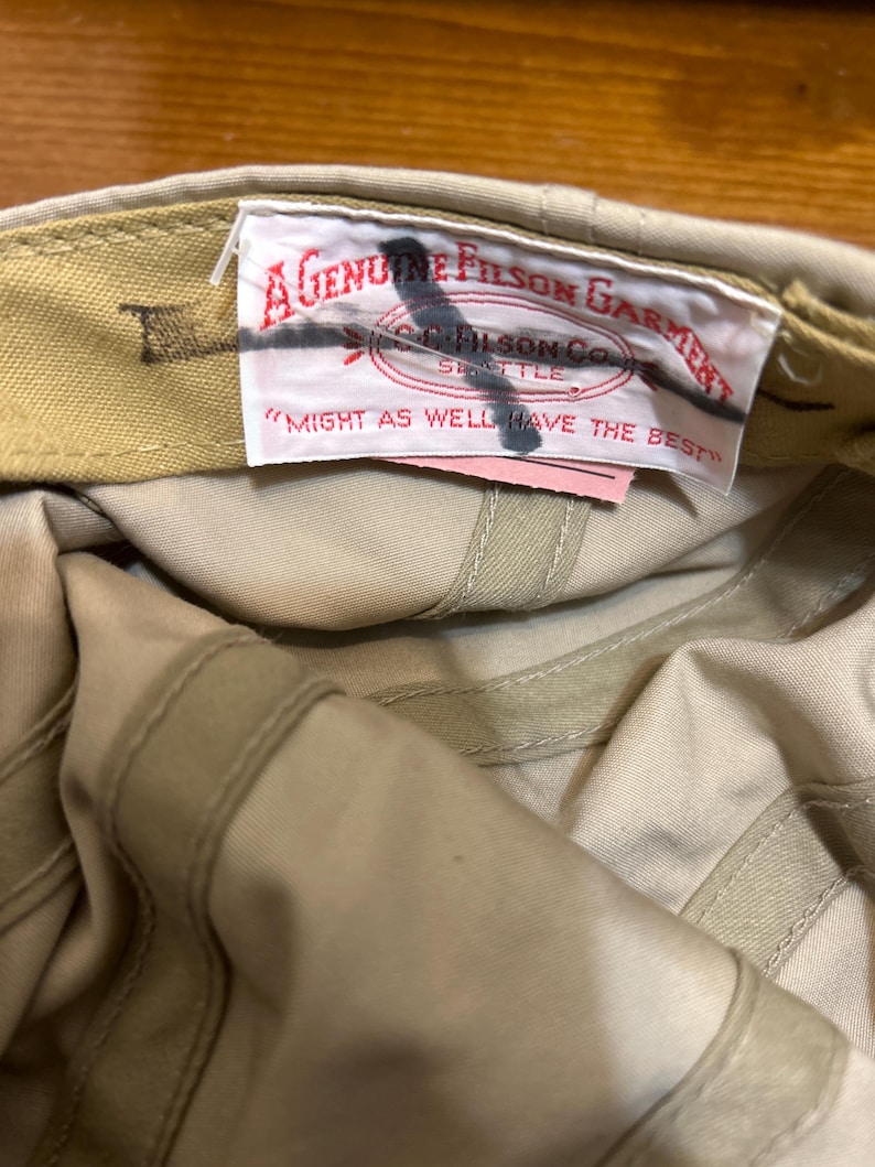 May include: Tan-colored cap with a label that reads "A Genuine Filson Garment" and "Might as well have the best." The cap is made of a durable fabric and features a classic design.