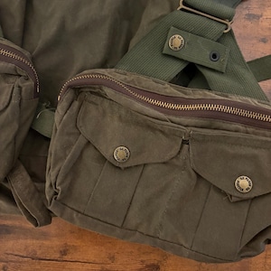 May include: Olive green canvas bag with multiple pockets and a shoulder strap. The bag features a zippered closure and brass-coloured hardware. The design includes two front pockets with button closures and a textured appearance.