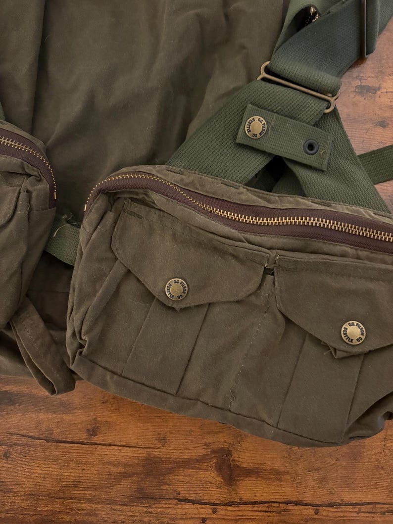 May include: Olive green canvas bag with multiple pockets and a shoulder strap. The bag features a zippered closure and brass-coloured hardware. The design includes two front pockets with button closures and a textured appearance.