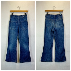 Puede incluir: Un par de pantalones vaqueros de campana azul de mezclilla con cierre de botón y dos bolsillos grandes en la parte delantera. Los pantalones vaqueros están colgados de una percha de madera.