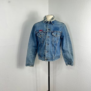 Può includere: Una giacca di jeans blu con un logo ricamato rosso sul petto sinistro. La giacca ha un colletto sfilacciato e un aspetto consumato.