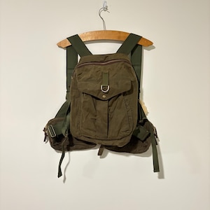 May include: An olive green backpack with multiple pockets and adjustable straps. The bag has a flap closure with a metal buckle and is suspended from a wooden hanger. The material appears to be durable, possibly water-resistant.