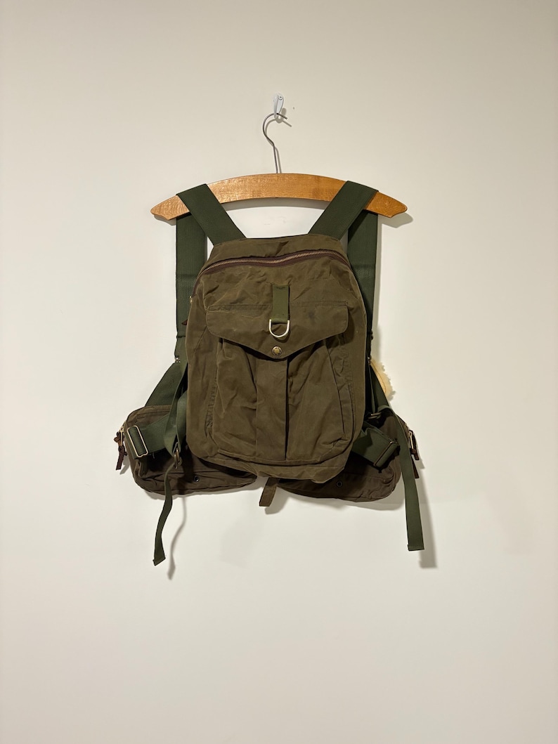 May include: An olive green backpack with multiple pockets and adjustable straps. The bag has a flap closure with a metal buckle and is suspended from a wooden hanger. The material appears to be durable, possibly water-resistant.