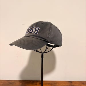 Peut inclure: Une casquette de baseball gris foncé avec le texte brodé 1969 en bleu marine et contour blanc sur le panneau avant. La casquette présente une visière incurvée avec des coutures visibles et une calotte structurée sur un support en fil noir.