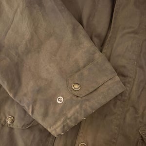 Peut inclure: Gros plan sur une manche de veste vert olive fonc&eacute;. La manche comporte un poignet boutonn&eacute; et plusieurs boutons m&eacute;talliques avec un motif circulaire. Le tissu a un aspect l&eacute;g&egrave;rement us&eacute; et textur&eacute;.