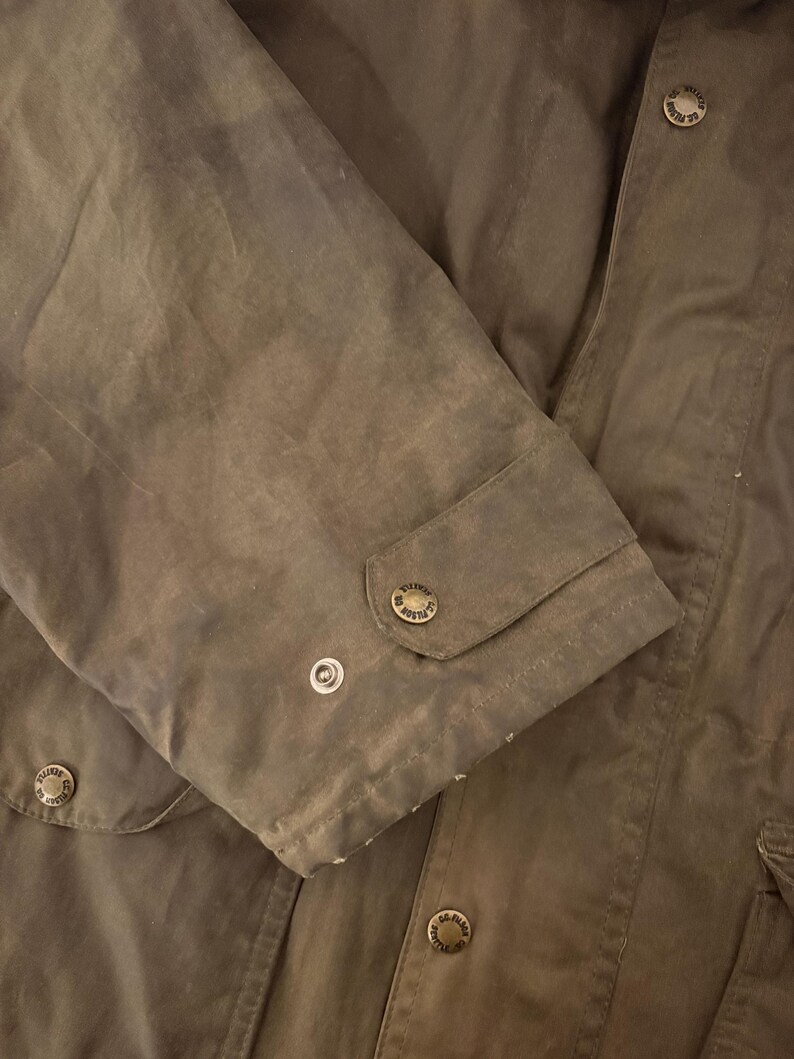 Peut inclure: Gros plan sur une manche de veste vert olive fonc&eacute;. La manche comporte un poignet boutonn&eacute; et plusieurs boutons m&eacute;talliques avec un motif circulaire. Le tissu a un aspect l&eacute;g&egrave;rement us&eacute; et textur&eacute;.