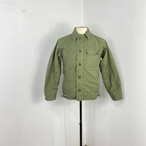 Puede incluir: Una chaqueta utilitaria verde con cierre de botones, un bolsillo en el pecho y mangas largas. La chaqueta tiene un corte holgado y está hecha de un tejido resistente.