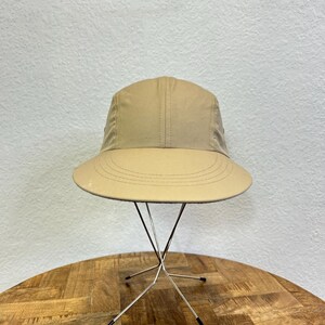 May include: A tan bucket hat with a wide brim. The hat is displayed on a silver metal stand with a starburst design. The hat has a structured crown and a stitched brim. The background is a neutral wall.