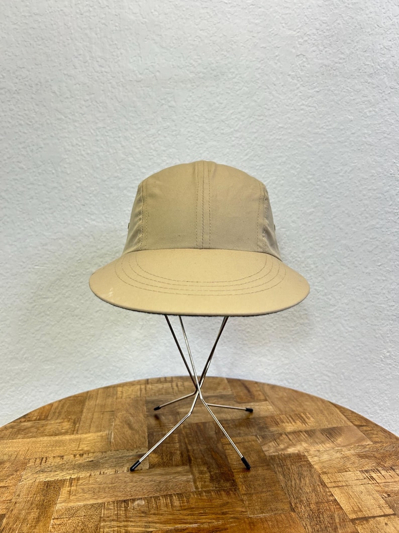 May include: A tan bucket hat with a wide brim. The hat is displayed on a silver metal stand with a starburst design. The hat has a structured crown and a stitched brim. The background is a neutral wall.