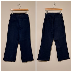 Può includere: Pantaloni in denim blu scuro con design a gamba larga. I pantaloni sono esposti su una gruccia di legno, mostrando le viste anteriore e posteriore. Presentano tasche anteriori e chiusura con bottoni.
