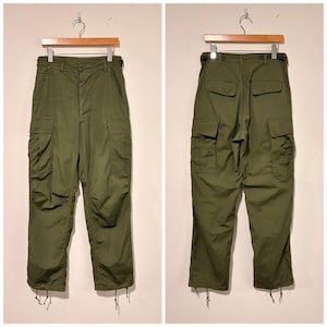 May include: Olive green cargo trousers featuring a button fly, two side cargo pockets with flaps, two back pockets with flaps, and adjustable drawstrings at the leg openings. The trousers are displayed on a wooden hanger, showing both front and back views.