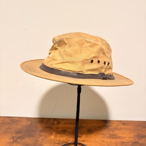 Pode incluir: Chap&eacute;u bege com aba larga e faixa de couro marrom escuro. O chap&eacute;u tem tr&ecirc;s pequenos orif&iacute;cios de ventila&ccedil;&atilde;o na lateral. O chap&eacute;u &eacute; exibido em um suporte de metal preto.
