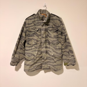 Puede incluir: Una chaqueta de camuflaje verde y gris con un patrón de rayas de tigre. La chaqueta tiene cuatro bolsillos delanteros, cierre de botones y cuello. La chaqueta está colgada de un gancho de metal.