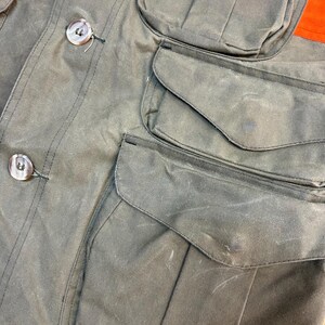 May include: A green waxed cotton jacket with multiple pockets. The jacket has a button closure and a contrasting orange panel on the top.