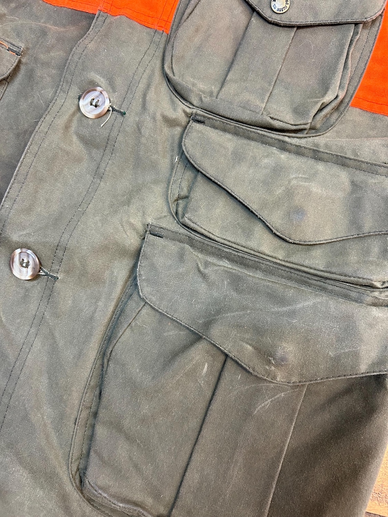 May include: A green waxed cotton jacket with multiple pockets. The jacket has a button closure and a contrasting orange panel on the top.