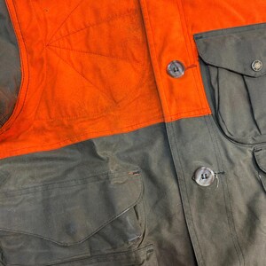 May include: A close-up of a brown and orange hunting jacket with multiple pockets and large buttons. The jacket has a contrasting orange panel on the upper chest.