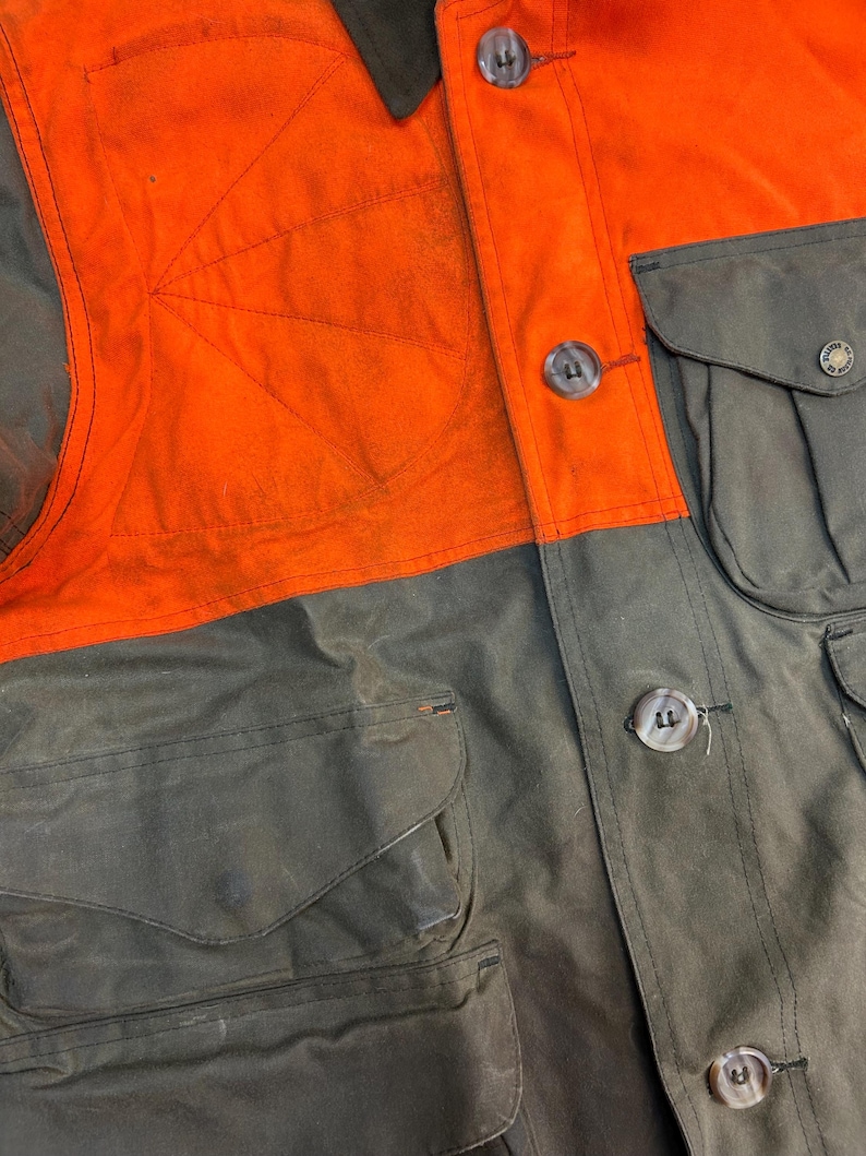 May include: A close-up of a brown and orange hunting jacket with multiple pockets and large buttons. The jacket has a contrasting orange panel on the upper chest.