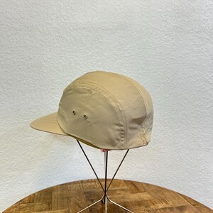 May include: A tan-colored, five-panel cap with a short brim. The hat is displayed on a metal stand with a wooden table in the foreground and a white wall in the background. The cap has two small ventilation eyelets on the side.