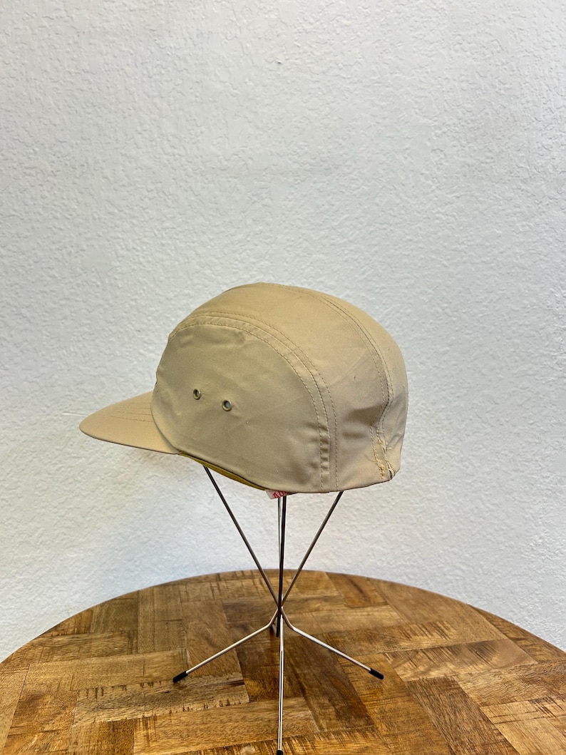 May include: A tan-colored, five-panel cap with a short brim. The hat is displayed on a metal stand with a wooden table in the foreground and a white wall in the background. The cap has two small ventilation eyelets on the side.
