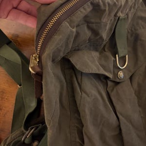 May include: An olive green backpack with a gold-coloured zip and hardware. The bag features adjustable green straps and a flap pocket. The material looks like waxed canvas. The bag is designed for carrying items, suitable for everyday use.