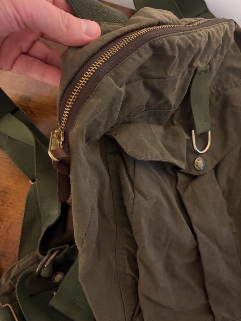 May include: An olive green backpack with a gold-coloured zip and hardware. The bag features adjustable green straps and a flap pocket. The material looks like waxed canvas. The bag is designed for carrying items, suitable for everyday use.