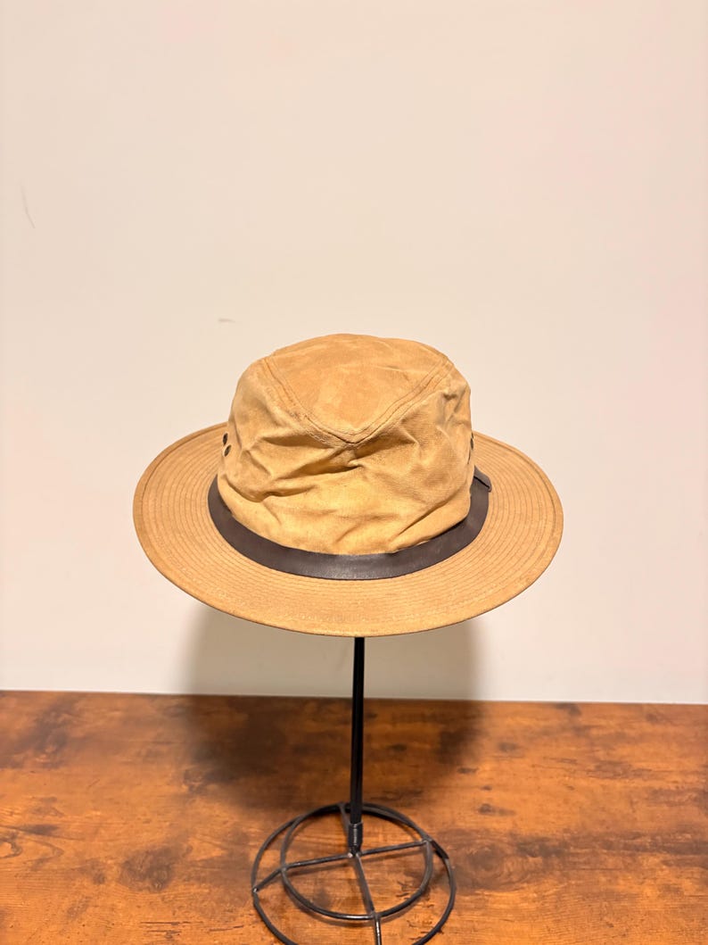 Pode incluir: Um chap&eacute;u bege com aba larga e uma faixa marrom escura. O chap&eacute;u tem uma apar&ecirc;ncia ligeiramente desgastada e &eacute; exibido em um suporte de metal preto. O chap&eacute;u parece ser feito de um material macio semelhante a camur&ccedil;a. O chap&eacute;u tem cerca de 30 cm de di&acirc;metro.