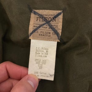May include: A close-up of a Filson brand tag, showing details of the product. The tag is cream-colored with black text, including the brand name, location, and product details. The tag is attached to a dark olive-green fabric.