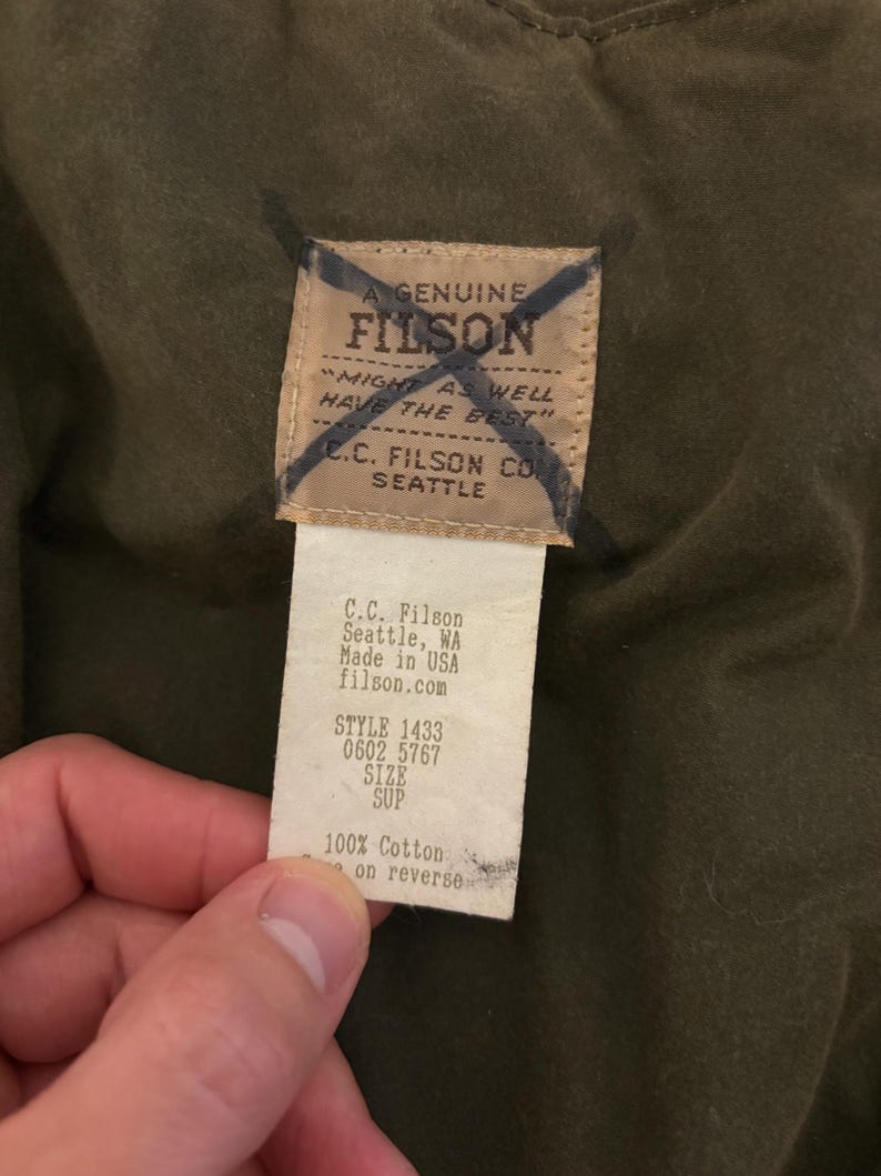 May include: A close-up of a Filson brand tag, showing details of the product. The tag is cream-colored with black text, including the brand name, location, and product details. The tag is attached to a dark olive-green fabric.